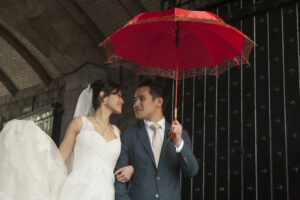 Bride and groom in Western style bridal outfits, gazing into each others eyes under a red umbrella