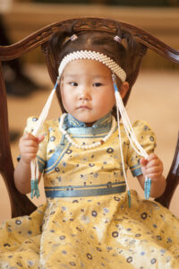 Little girl wearing gold and blue Chinese outfit, holding on to tassels on her head piece