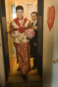 Groom exiting a room wearing gold and red traditional Chinese outfit, holding red envelopes and a pink rose bouquet