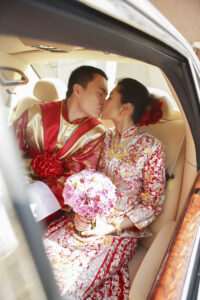 bride and groom in traditional red and gold Chinese wedding attire kissing in car. Bride is holding pink flowers