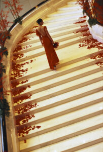 View of the back of a bride in a sleek red dress walking up a cream marble staircase with rose petals cascading either side, carrying a red rose bouquet