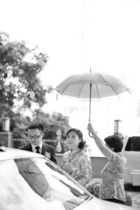 black and white image of lady holding umbrella over bridal party whilst they are getting in a car