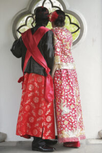 Bride and Groom wearing traditional black, red and gold Chinese wedding outfits looking through a window